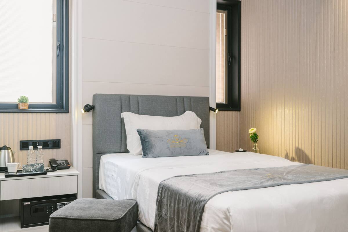 Habitació d'hotel ben ordenada amb un llit, una taula de nit i una finestra petita amb una planta.