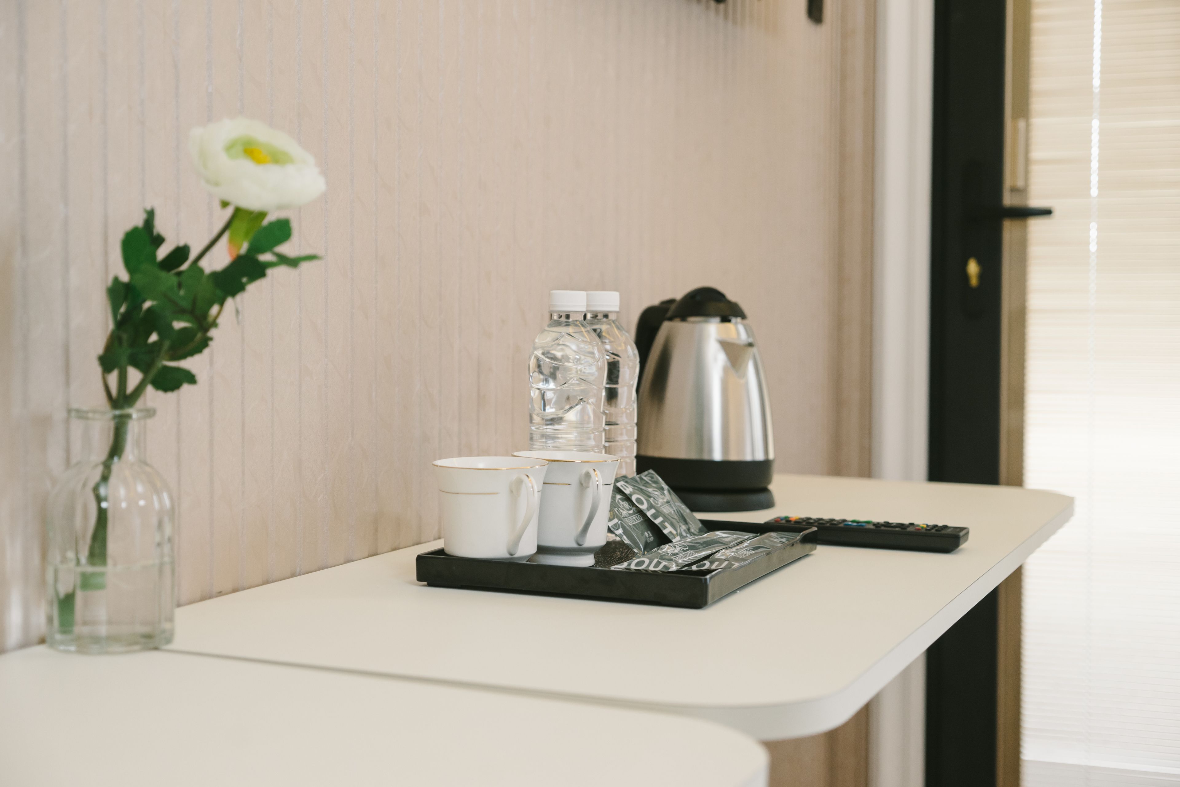 Una mesa blanca con un jarrón de vidrio transparente que contiene una flor blanca, dos tazas blancas, una bandeja negra con un control remoto, una botella de vidrio y una tetera plateada.