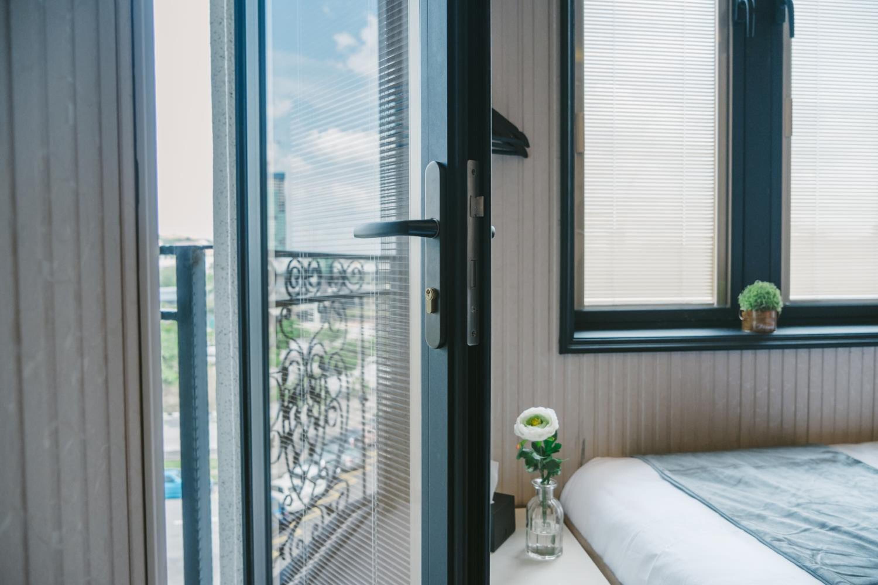 A bedroom with a bed, a window with blinds, a small vase with a flower, and a view of the outside balcony.