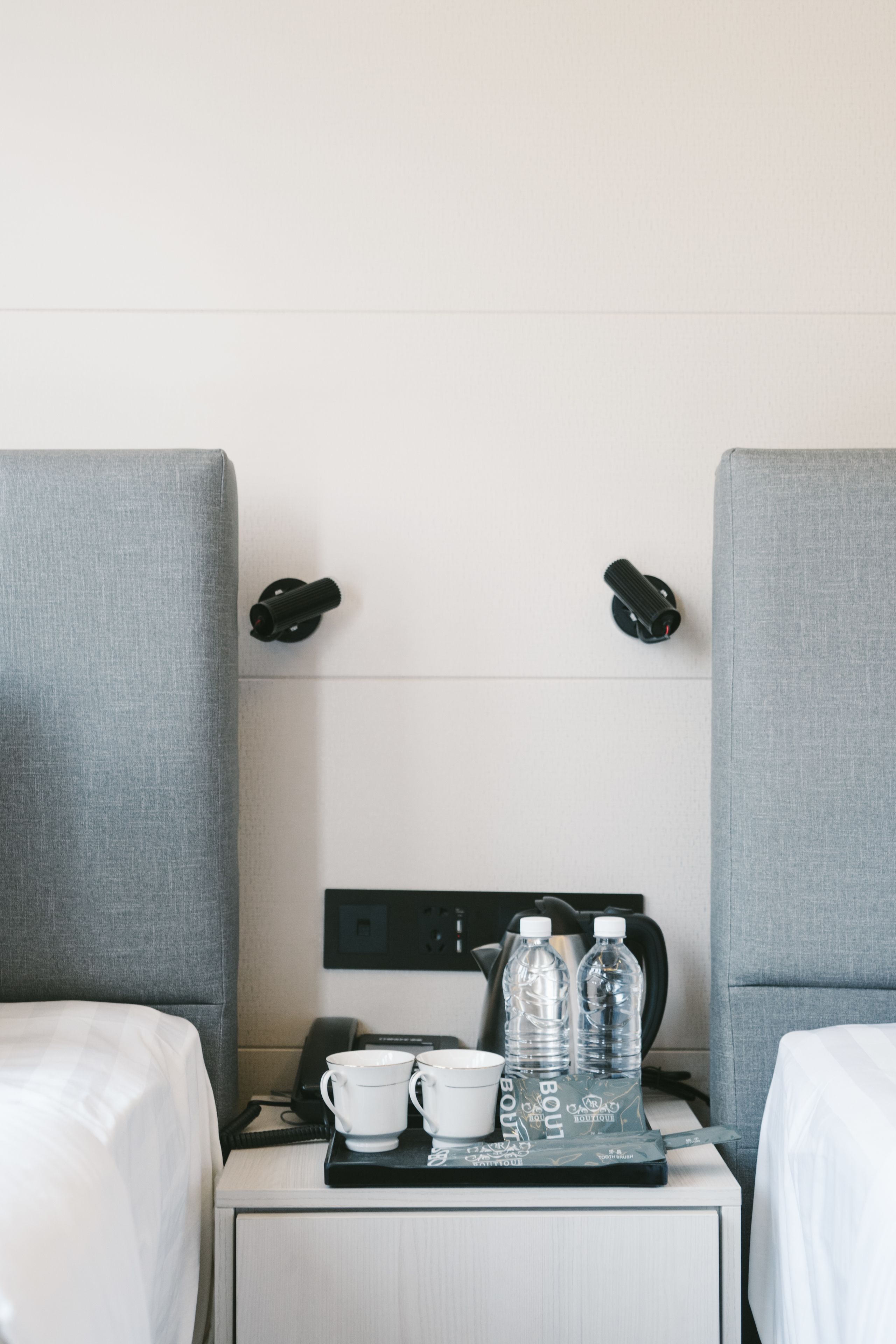 A hotel room with two single beds, a small bedside table with a phone, a tray with cups and water bottles, and two wall-mounted lamps.
