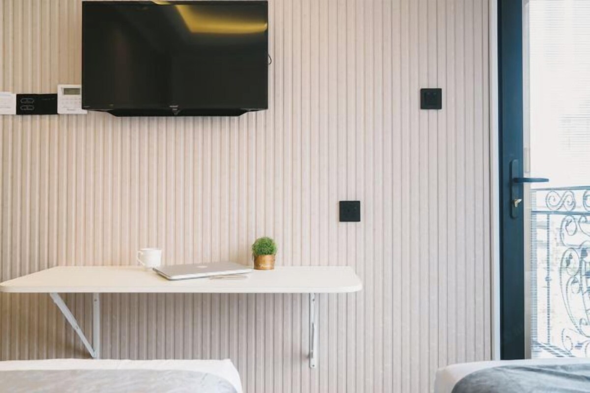 Una camera d'albergo con un letto, una scrivania con un portatile, una piccola pianta e una TV a muro.