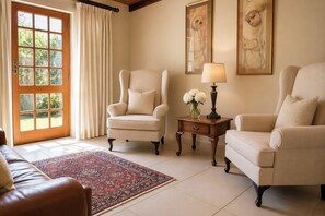 A room with two armchairs, a small table with a lamp and flowers, a patterned rug, and a large window with curtains. - Bakenskraal Ostrich Palace (Oudtshoorn)