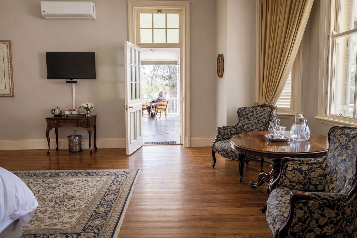 A room with a wooden floor, a round table with chairs, a television on the wall, and a door leading to an outdoor area.