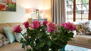 Ein Wohnzimmer mit Rosenblumen in einer Vase, einer Couch mit floralen Kissen und einer Lampe auf einem Beistelltisch.