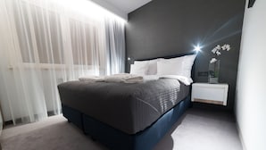 A modern bedroom with a dark blue bed, white pillows, and a bedside table with a vase of flowers. - Apart-Hotel VIVI Residence & SPA (Zory)