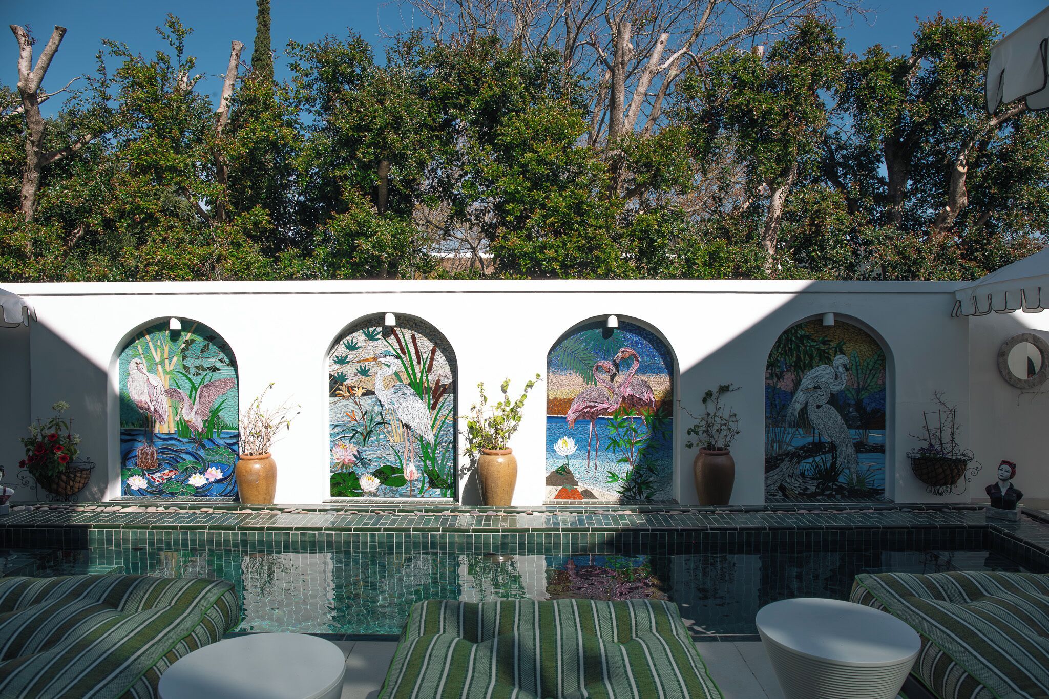 Uma área de piscina com piso de azulejos, um espaço de estar com almofadas listradas e quatro janelas em arco com trabalhos em mosaico.