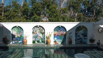 Uma área de piscina com piso de azulejos, um espaço de estar com almofadas listradas e quatro janelas em arco com trabalhos em mosaico.