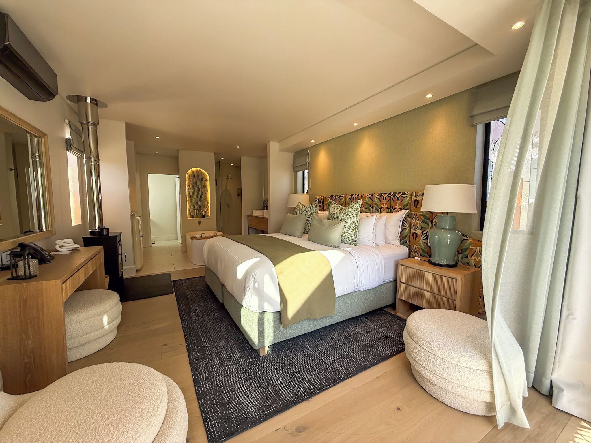 Habitación de hotel moderna con una cama grande, mesitas de noche, un escritorio, un espejo y un baño visible a través de una puerta abierta.