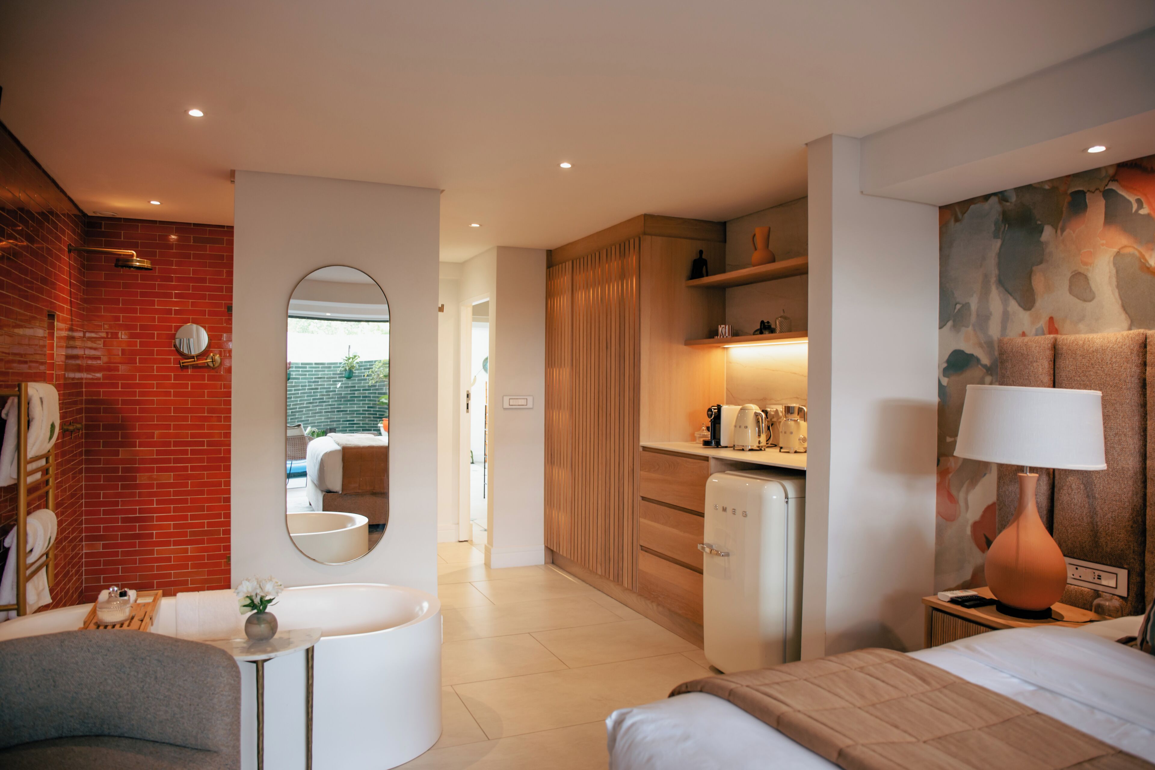 Una habitación de hotel moderna con pared de ladrillo, un espejo redondo, una mesita con un florero, una cama con cabecera, una mesita de noche con lámpara y una zona de kitchenette.