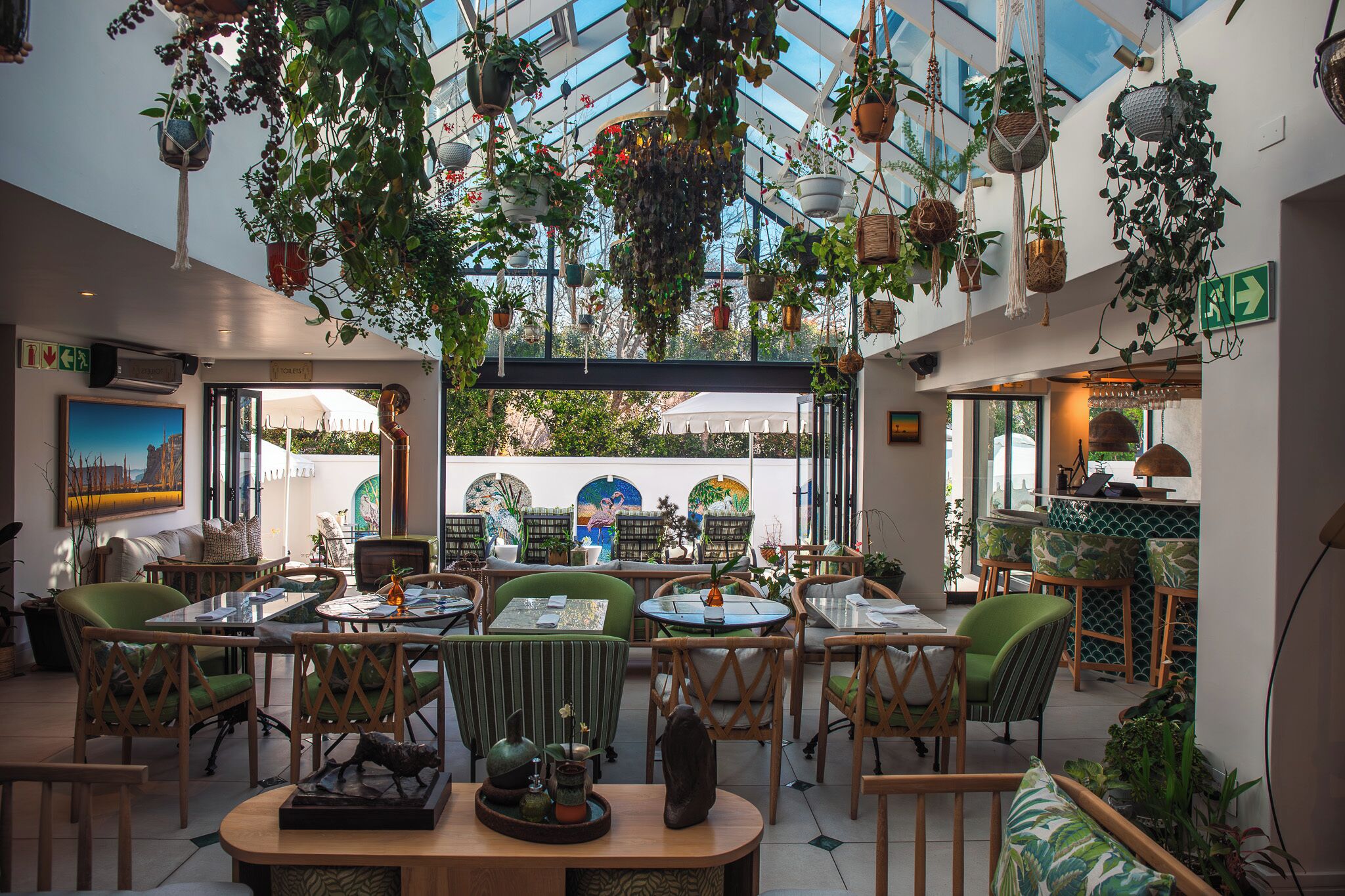 Een restaurant met binnenzitplaatsen, hangplanten en uitzicht door grote ramen.