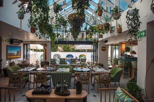 Un restaurante con mesas dentro, plantas colgantes y vistas amplias a través de grandes ventanales.