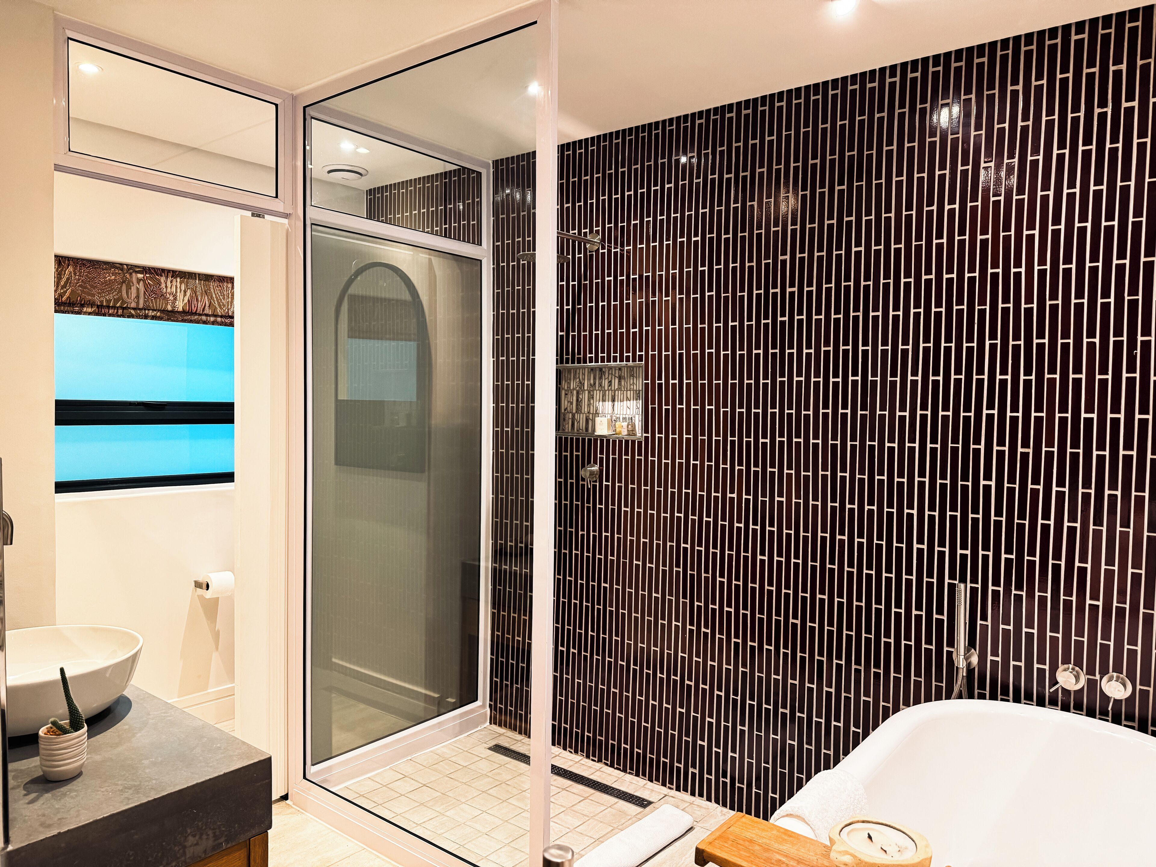 Un baño moderno con mampara de ducha de vidrio, un lavabo blanco con cuenco y pared revestida de baldosas negras.