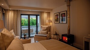 Habitación de hotel con cama, una silla, una mesa con una botella y vasos, una chimenea y vistas a zonas verdes por la ventana.