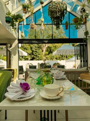 An outdoor dining area with a table set for two, surrounded by hanging plants and a view of the outside.