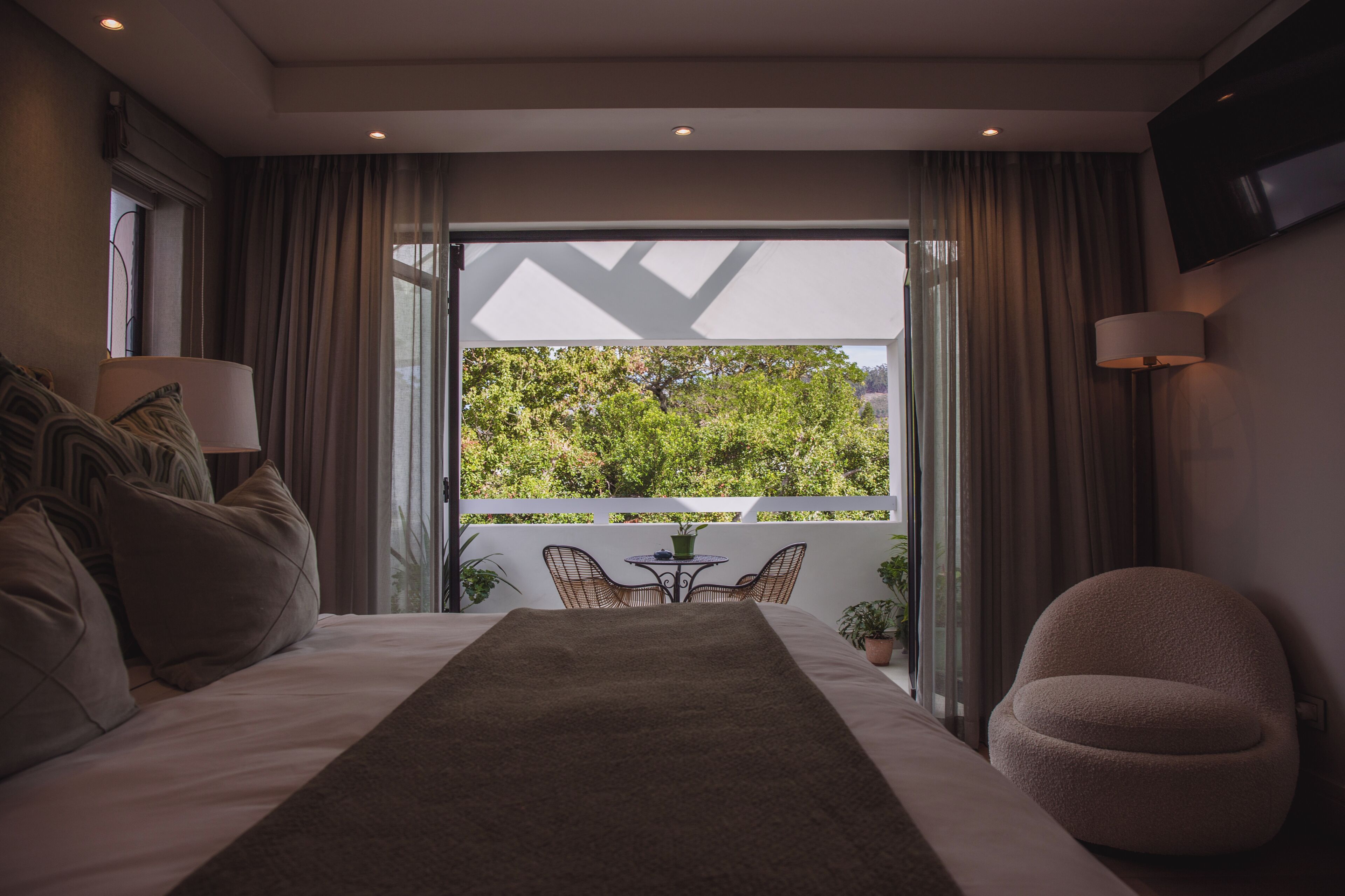 Quarto com uma cama grande, uma mesinha com cadeiras, televisão na parede e vista para o verde lá fora.