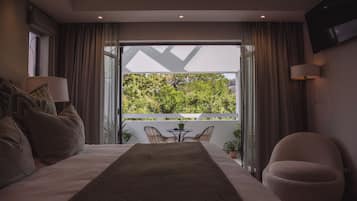 Quarto com uma cama grande, uma mesinha com cadeiras, televisão na parede e vista para o verde lá fora.