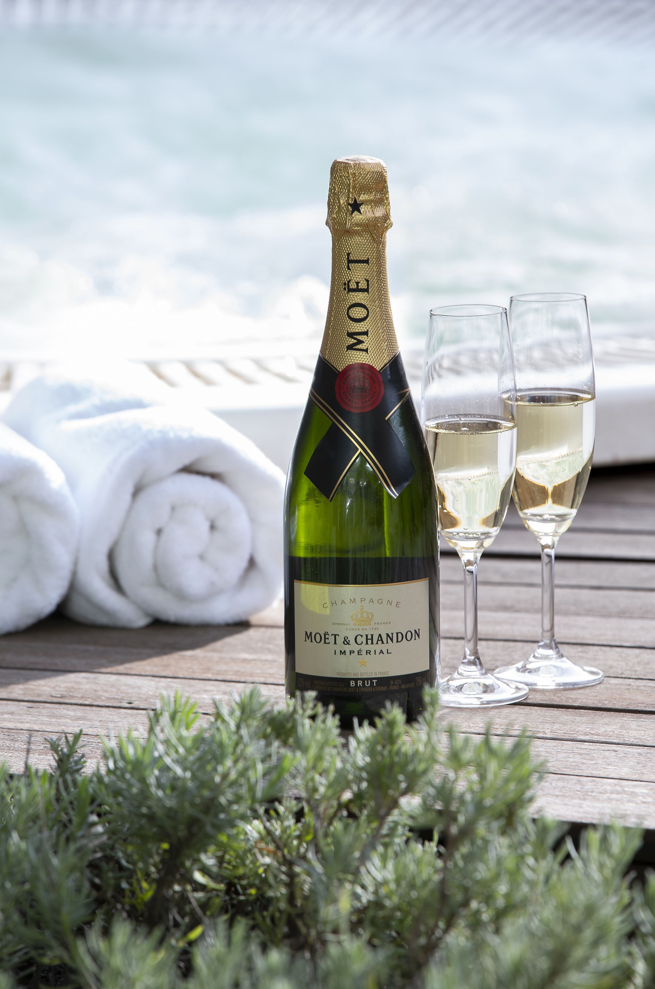 En flaske Moët & Chandon Brut champagne, to glas og et træterrasse med en pool i baggrunden.