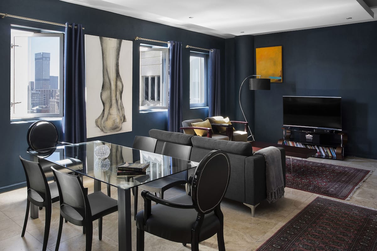 Een moderne woonkamer met een glazen eettafel, zwarte stoelen, een grijze bank, een flatscreen-tv en een boekenkast.