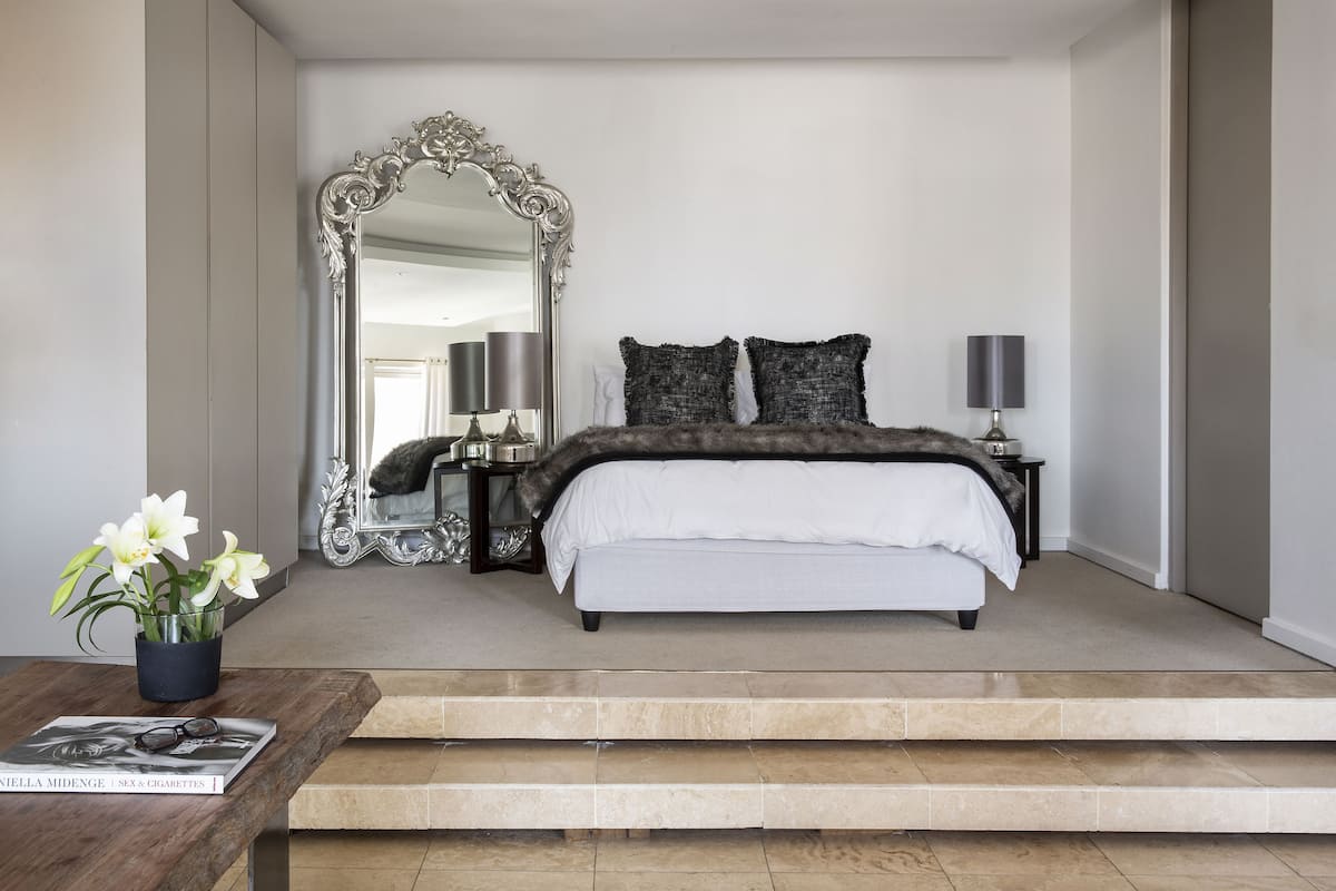 Een slaapkamer met een grote, versierde spiegel, een bed met donkere beddengoed, twee bedlampjes en een bijzettafeltje met een tijdschrift.