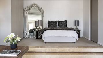 A bedroom with a large ornate mirror, a bed with dark bedding, two bedside lamps, and a coffee table with a magazine.