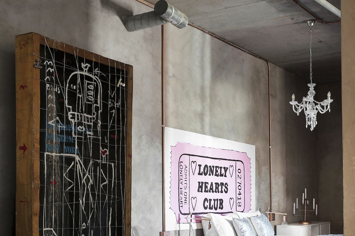 Een slaapkamer met een bed, nachtkastje, dressoir, een kroonluchter en een muur met een bord waarop 'Lonely Hearts Club' staat.