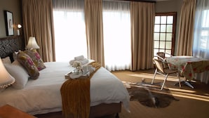 A hotel room with a large bed, a table with a floral tablecloth, a chair, a lamp, and a window with curtains.