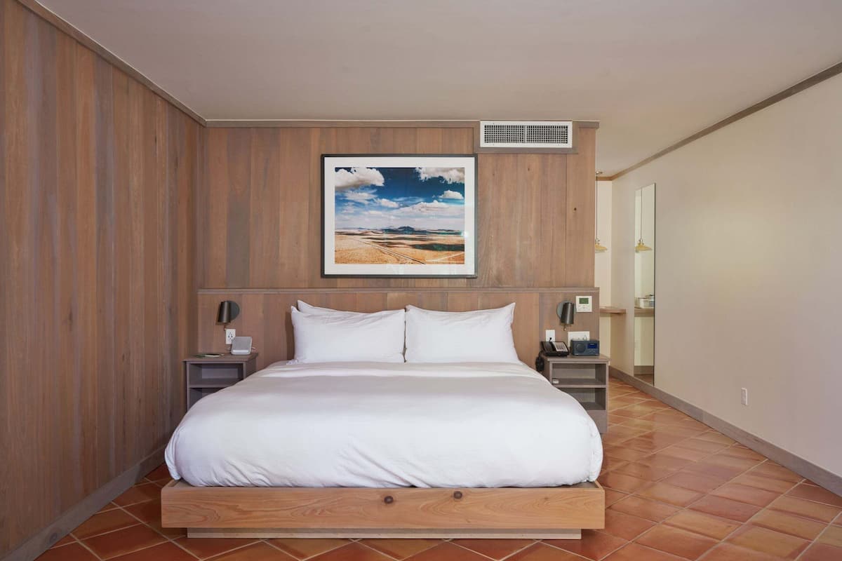 A bedroom with a wooden headboard, a large bed, two bedside tables with lamps, a wall-mounted air vent, and a framed picture above the bed.
