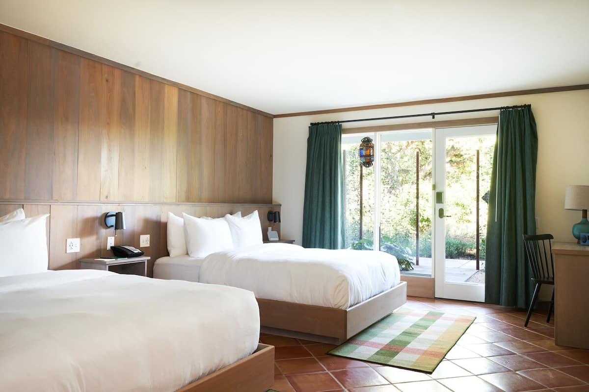 A hotel room with two beds, a wooden headboard, a nightstand with a phone, a desk with a lamp, and a view of the outdoors through a sliding glass door.