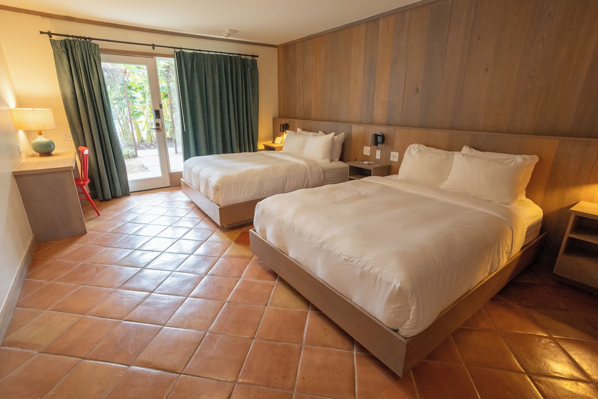 A hotel room with two beds, a wooden headboard, a nightstand, a lamp, and a view through a glass door.