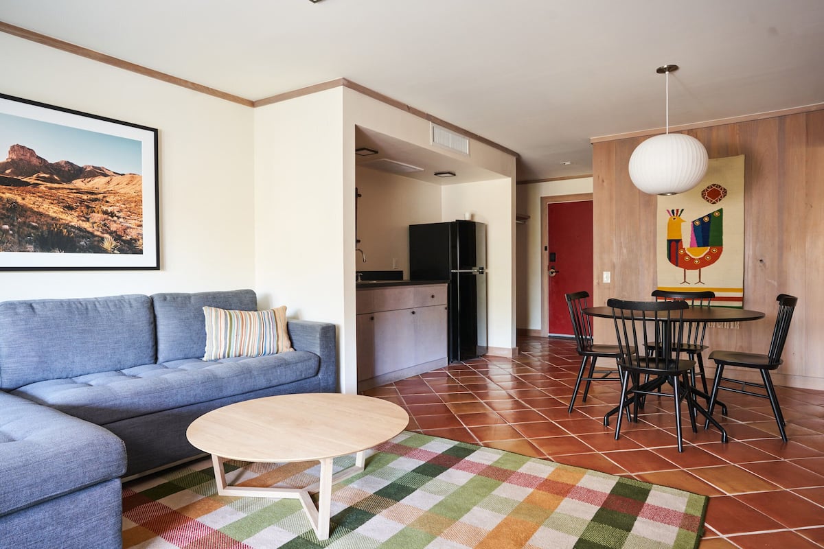 A living room with a sofa, a round coffee table, and a colorful rug. There is a dining area with a table and chairs. A kitchen is visible in the background.