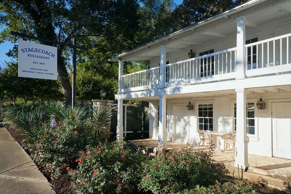 A white two-story building with a covered porch, a signboard, and a garden with red flowers.