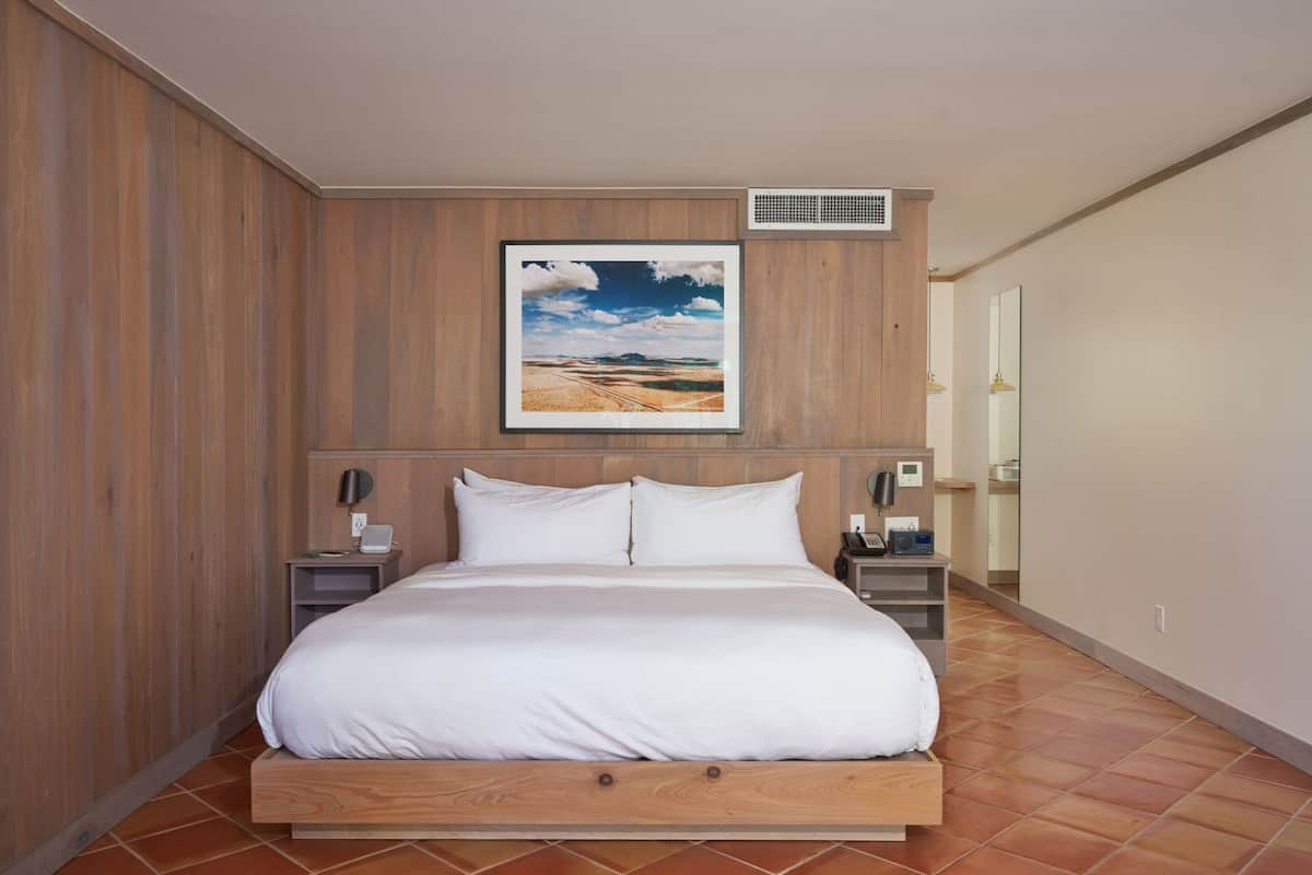 A bedroom with a wooden bed, a nightstand with a phone and a lamp, a wall-mounted air vent, and a framed picture above the bed.