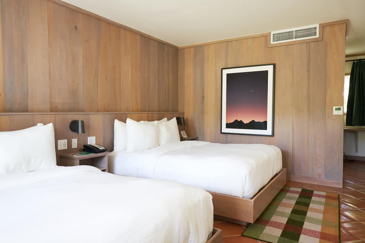A hotel room with two beds, a nightstand with a phone, a framed picture on the wall, and a check-in station visible through a doorway.