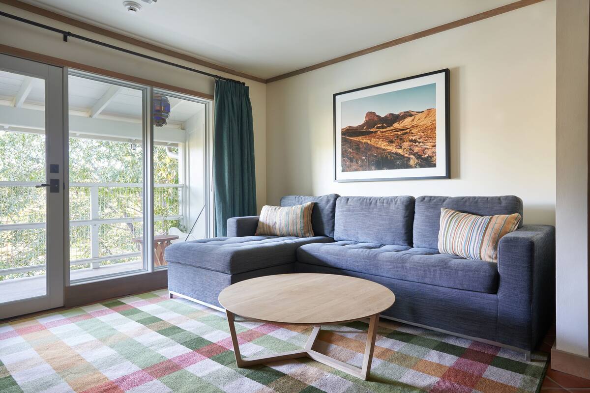 A living room with a sectional sofa, a round coffee table, a checkered rug, and a large window with a view of trees and mountains.