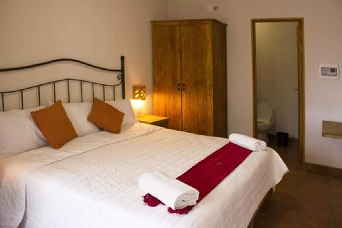 Un lit bien fait, avec du linge de lit blanc et des oreillers marron, une armoire en bois et une salle de bain visible à travers une porte ouverte.