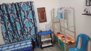 Une chambre à coucher avec un lit, une chaise bleue, une fenêtre avec un rideau, une étagère avec des vêtements et une horloge murale.