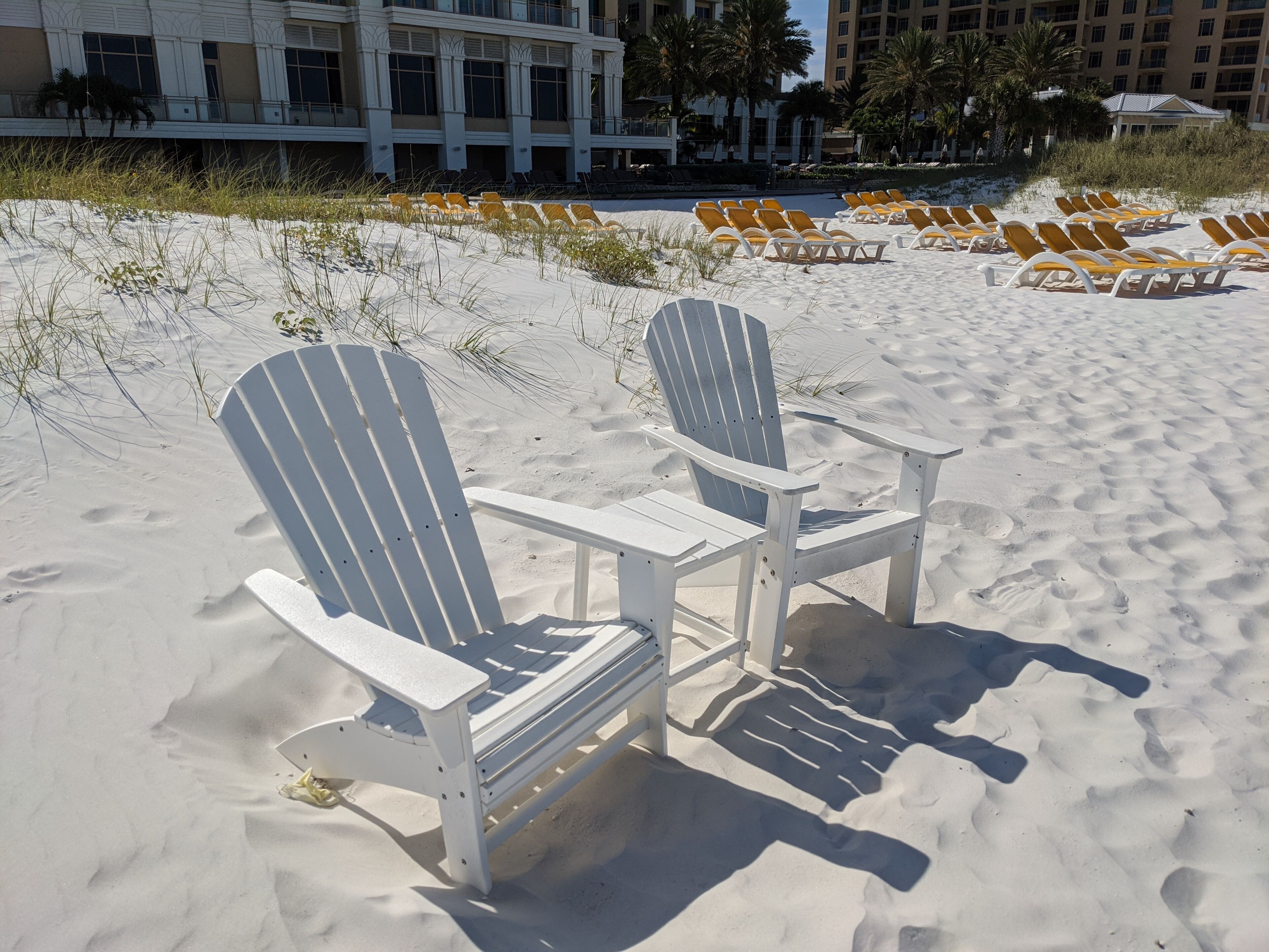 A sandy beach with white Adirondack chairs and yellow lounge chairs.
