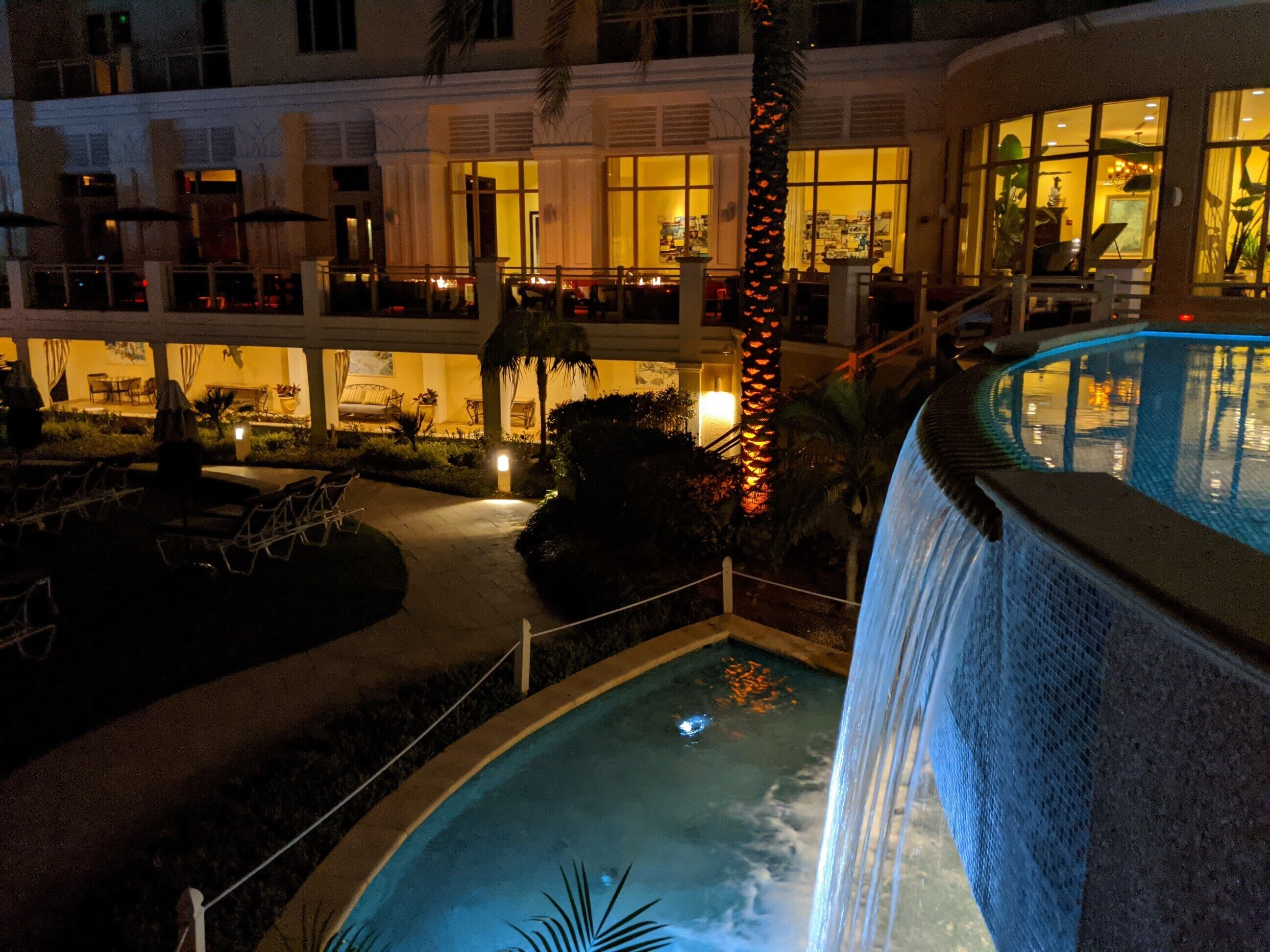 A nighttime view of a hotel with a pool and waterfall. The pool area is illuminated, and there are lounge chairs and a palm tree visible.