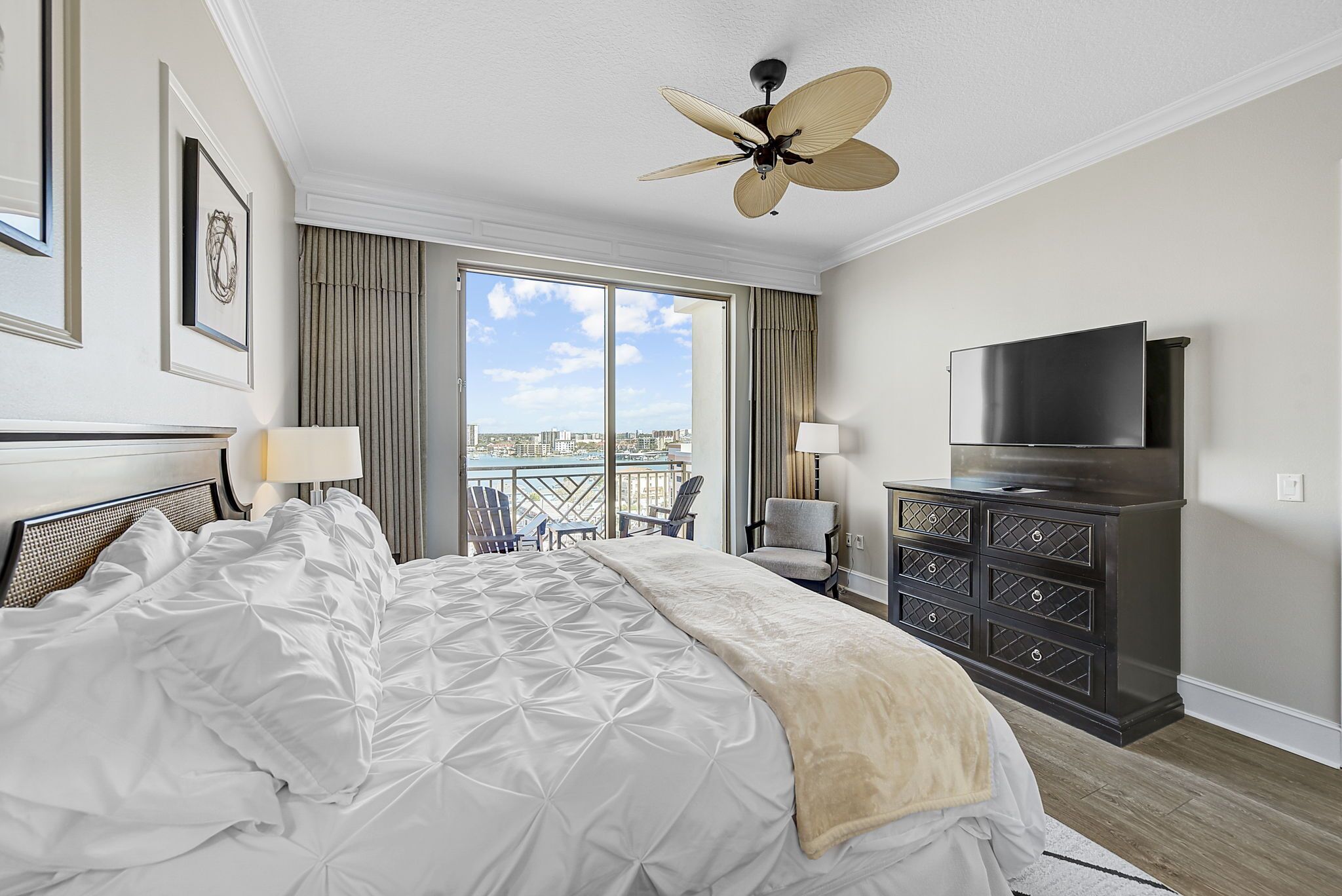 A hotel room with a large bed, a ceiling fan, a TV, and a view of the city through the window.