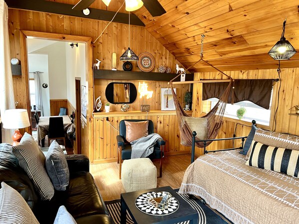 A cozy cabin interior with a wooden ceiling, a bed with patterned bedding, a dining area, and a hammock.