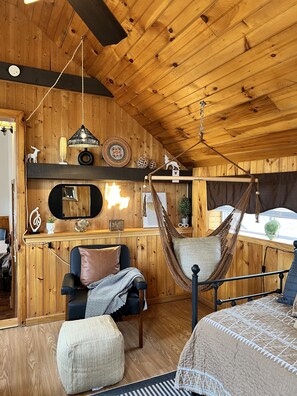 A cozy room with wooden walls, a hammock, a chair, a bed, and a mirror.