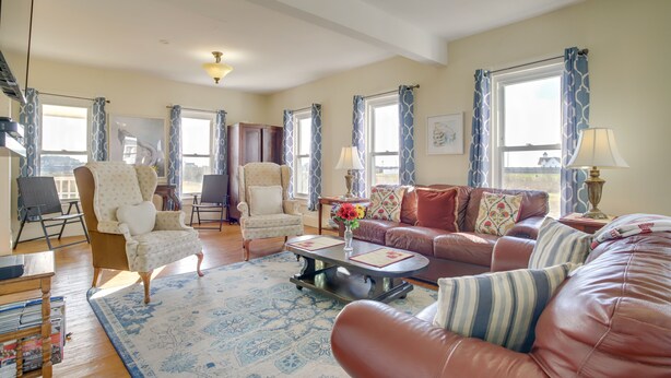 A spacious living room with a large sofa, armchairs, and a coffee table. There are multiple windows with curtains, a chandelier, and a rug.