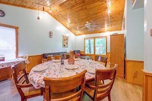 Un comedor con una mesa redonda preparada para cuatro personas, sillas de madera, un ventilador de techo y una ventana con vista a árboles.