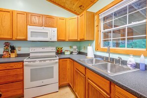 Cocina con armarios de madera, microondas, estufa, fregadero y ventana con vista a follaje.