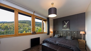 A bedroom with a large window, a bed with a dark quilt, two bedside tables with lamps, and a wall-mounted light fixture. - Beautiful location with indoor swimming pool, wellness area & breathtaking view (Ventron)