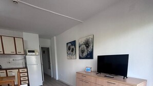 Eine moderne Küche mit weißen Schränken, einer Mikrowelle und einem Kühlschrank. Ein Essbereich mit Holztisch und Stühlen lädt zum Verweilen ein. Ein Fernseher steht auf einer Holzkommode, und an der Wand befinden sich zwei gerahmte Kunstwerke.