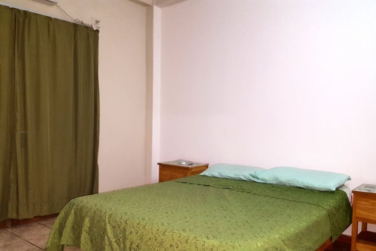 A small, simple bedroom with a single bed, a wooden nightstand, a green curtain, and a plain white wall.