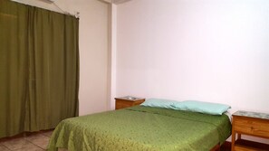Um quarto pequeno e simples, com uma cama de solteiro, uma mesa de cabeceira de madeira, uma cortina verde e uma parede branca lisa.