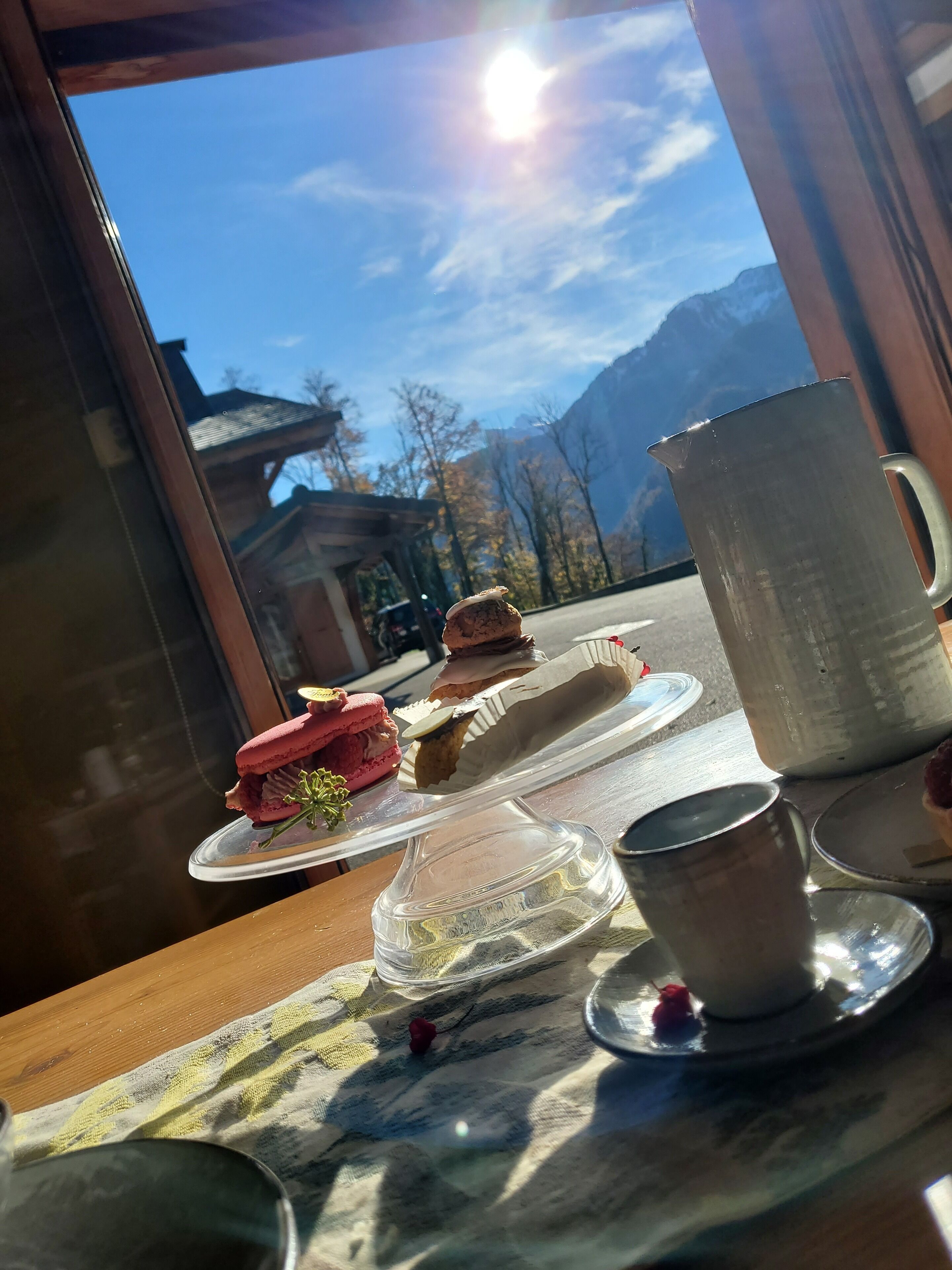 Una mesa con vista despejada a un paisaje montañoso a través de una ventana, una jarra metálica y un plato con alimentos.