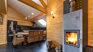 A cozy cabin interior with a stone fireplace, wooden walls, a dining area with wicker chairs, and a kitchen with a modern refrigerator. - Vacation Rental for 10 People: 4 Bedrooms, Indoor Pool, Sauna, Fitness Center in Ventron, Vosges (Ventron)